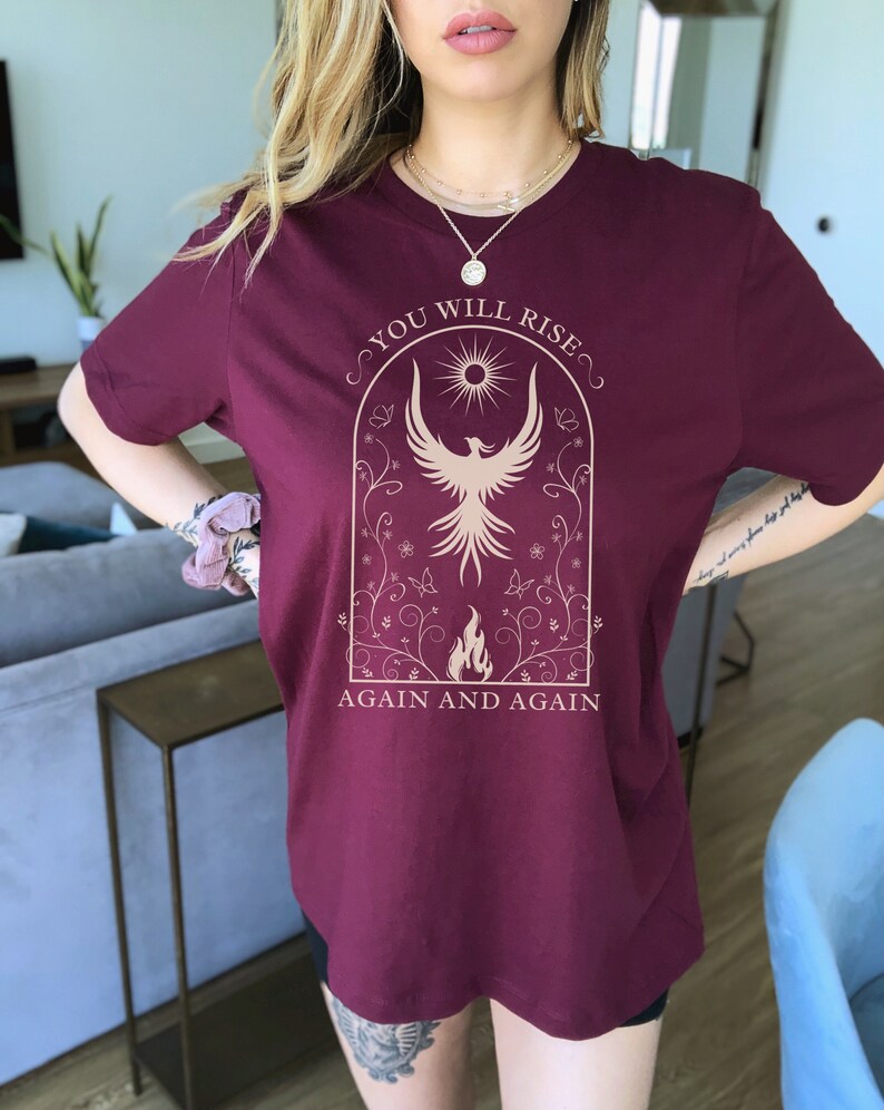 May include: A maroon t-shirt with a graphic of a phoenix rising from flames in a floral arch. The text "You Will Rise Again and Again" is printed below the graphic.