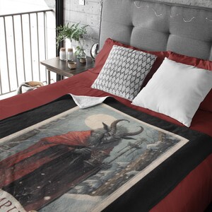 May include: A black and white throw blanket with a vintage illustration of a horned figure in a red cloak holding a cup. The illustration is set against a snowy background with a full moon. The text "Krampus" is visible at the bottom of the illustration.