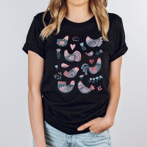 K&ouml;nnte beinhalten: Schwarzes T-Shirt mit einem bunten Muster aus stilisierten V&ouml;geln und Blumen. Die V&ouml;gel sind rosa, blau und grau mit filigranen Details.