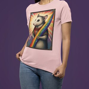 Pode incluir: Uma camiseta rosa com um gr&aacute;fico de um gamb&aacute; segurando uma bandeira arco-&iacute;ris. O gamb&aacute; est&aacute; usando um colar de ouro e uma capa vermelha. O fundo &eacute; um arco-&iacute;ris com estrelas.