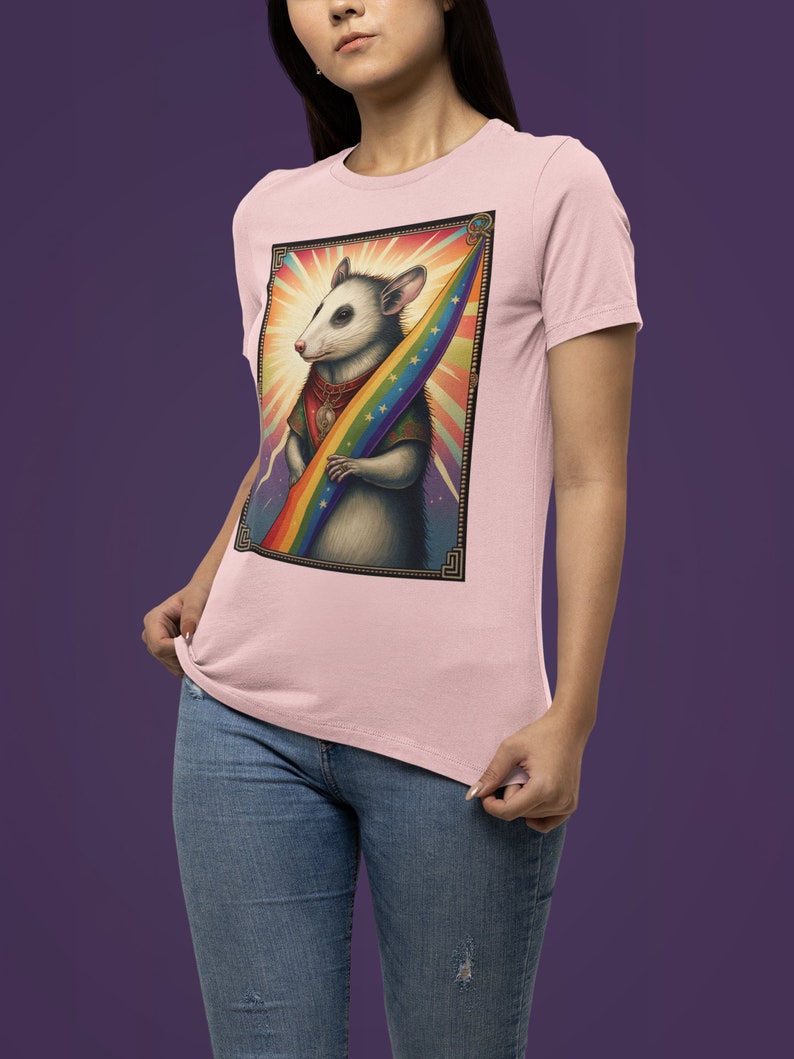 Pode incluir: Uma camiseta rosa com um gr&aacute;fico de um gamb&aacute; segurando uma bandeira arco-&iacute;ris. O gamb&aacute; est&aacute; usando um colar de ouro e uma capa vermelha. O fundo &eacute; um arco-&iacute;ris com estrelas.