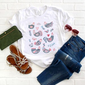 K&ouml;nnte beinhalten: Ein wei&szlig;es T-Shirt mit einem bunten Muster aus stilisierten H&uuml;hnern und floralen Designs. Das Shirt liegt auf einem wei&szlig;en Ziegelhintergrund mit einer blauen Jeans, einer gr&uuml;nen Brieftasche und braunen Sandalen.