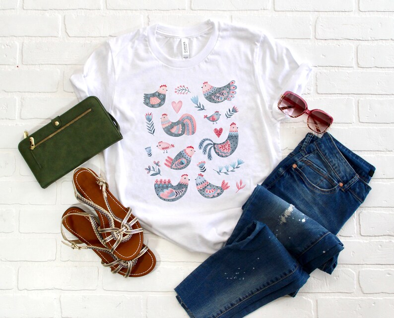 K&ouml;nnte beinhalten: Ein wei&szlig;es T-Shirt mit einem bunten Muster aus stilisierten H&uuml;hnern und floralen Designs. Das Shirt liegt auf einem wei&szlig;en Ziegelhintergrund mit einer blauen Jeans, einer gr&uuml;nen Brieftasche und braunen Sandalen.