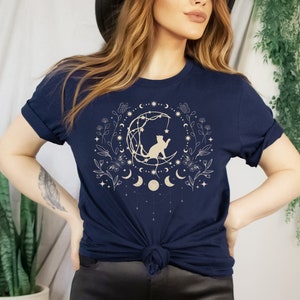 May include: A navy blue t-shirt with a gold graphic of a cat sitting on a crescent moon surrounded by stars, flowers, and moons.