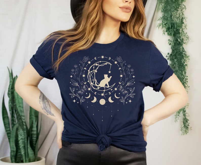 May include: A navy blue t-shirt with a gold graphic of a cat sitting on a crescent moon surrounded by stars, flowers, and moons.