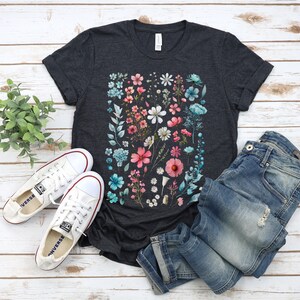 May include: A dark grey t-shirt with a floral print featuring pink, blue, and white flowers. The shirt is laid flat on a white wooden surface with a pair of white sneakers and blue jeans.