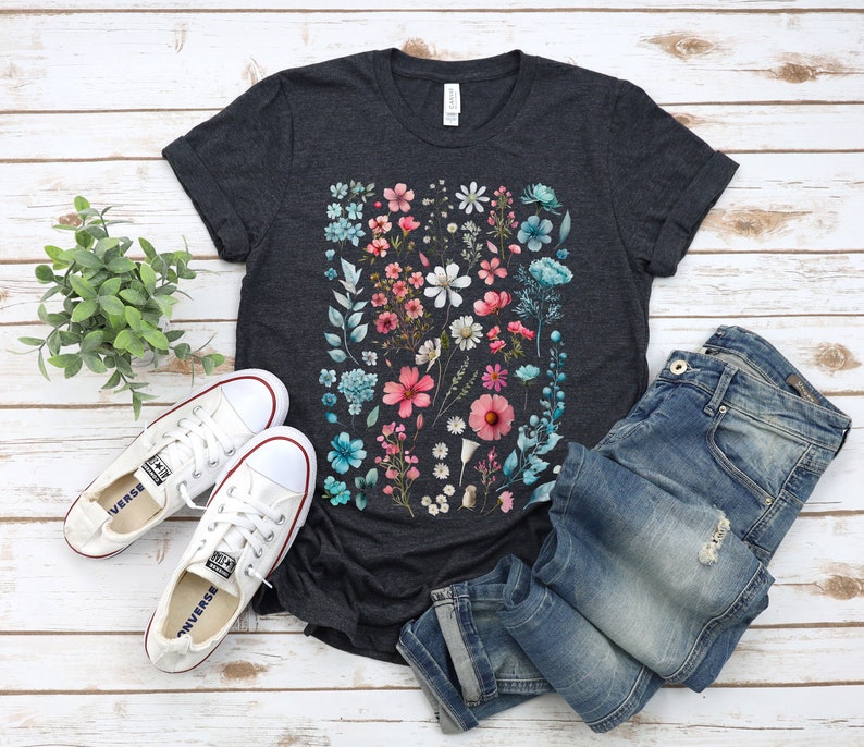 May include: A dark grey t-shirt with a floral print featuring pink, blue, and white flowers. The shirt is laid flat on a white wooden surface with a pair of white sneakers and blue jeans.