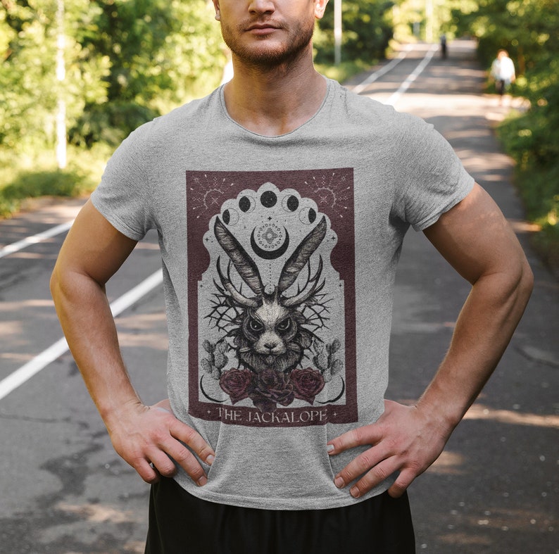 K&ouml;nnte beinhalten: Hellgraues T-Shirt mit einer detaillierten Illustration eines Jackalopen mit Geweih, Rosen und einem Mondphasen-Design. Der Text "THE JACKALOPE" ist unter dem Bild aufgedruckt. Das Shirt besteht aus einem weichen Material.