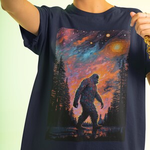 May include: A navy blue t-shirt with a graphic print of a Bigfoot walking through a forest at sunset. The sky is a vibrant mix of orange, pink, and blue with stars and planets.