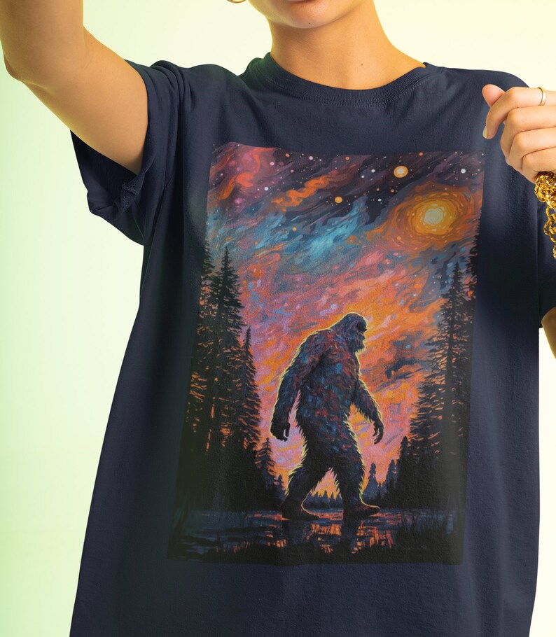 May include: A navy blue t-shirt with a graphic print of a Bigfoot walking through a forest at sunset. The sky is a vibrant mix of orange, pink, and blue with stars and planets.