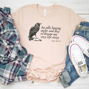 May include: A peach-coloured t-shirt featuring a black and white illustration of a raven and a quote from Edgar Allan Poe: "An idle longing night and day to dream my very life away".