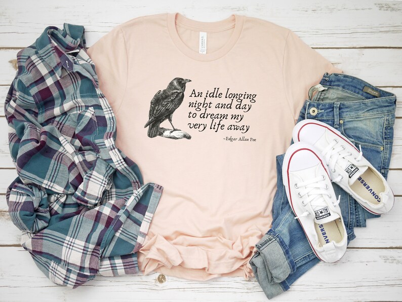 May include: A peach-coloured t-shirt featuring a black and white illustration of a raven and a quote from Edgar Allan Poe: "An idle longing night and day to dream my very life away".