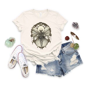 May include: A beige t-shirt with a graphic print of a skeleton fairy with wings. The fairy is surrounded by a decorative frame.