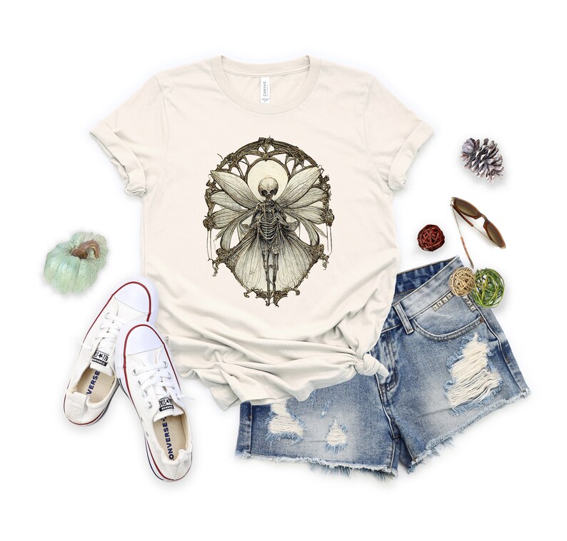 May include: A beige t-shirt with a graphic print of a skeleton fairy with wings. The fairy is surrounded by a decorative frame.