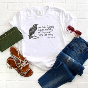May include: White t-shirt featuring a black and white illustration of a raven and a quote from Edgar Allan Poe: "An idle longing night and day to dream my very life away". The t-shirt is styled with blue jeans, a green clutch bag, and brown sandals.