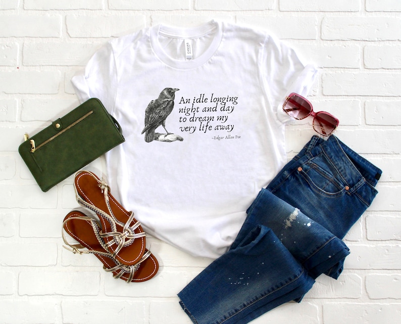 May include: White t-shirt featuring a black and white illustration of a raven and a quote from Edgar Allan Poe: "An idle longing night and day to dream my very life away". The t-shirt is styled with blue jeans, a green clutch bag, and brown sandals.