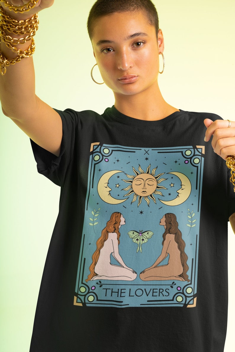 May include: Black t-shirt with a graphic print of a tarot card depicting two figures, a sun, a moon, and a moth. The card is labeled "THE LOVERS".