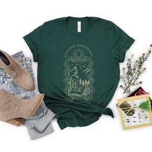 K&ouml;nnte beinhalten: Ein dunkelgr&uuml;nes T-Shirt mit einem goldenen Grafikdesign aus einer Bergkette, einem Buch und einem Halbmond. Der Text "Fictional Wandererlust" ist in einer goldenen Schriftart um die Grafik herum geschrieben.