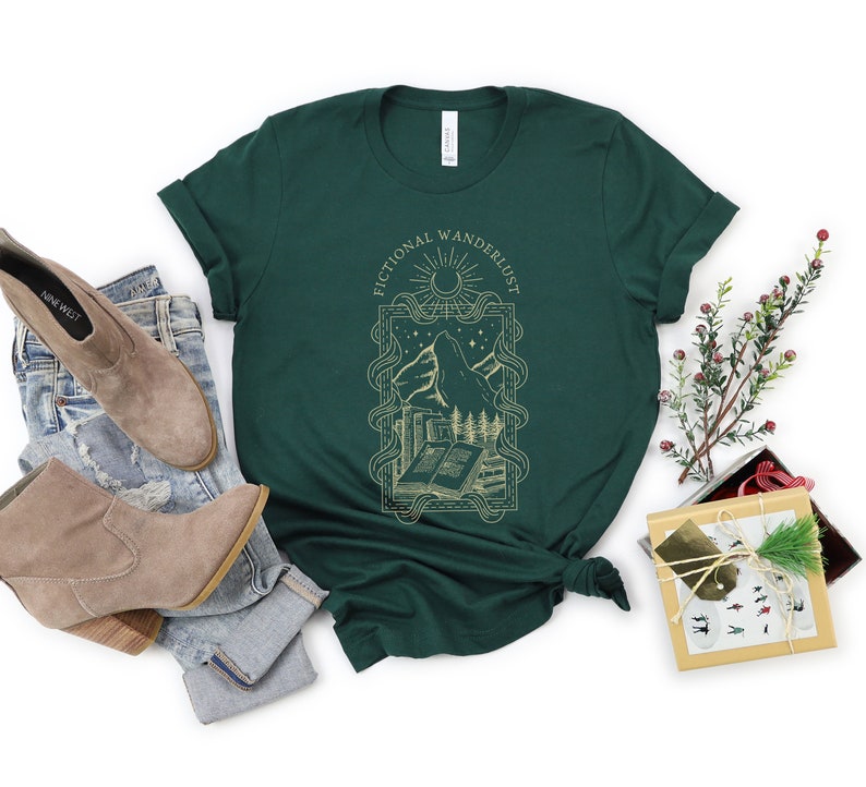 K&ouml;nnte beinhalten: Ein dunkelgr&uuml;nes T-Shirt mit einem goldenen Grafikdesign aus einer Bergkette, einem Buch und einem Halbmond. Der Text "Fictional Wandererlust" ist in einer goldenen Schriftart um die Grafik herum geschrieben.
