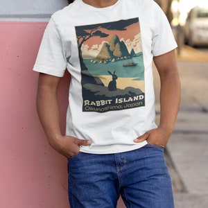 May include: White t-shirt with a vintage-style graphic of a rabbit island in Japan. The graphic features a small rabbit sitting on a rock, a boat in the water, and a mountain range in the background. The text "Rabbit Island" and "Okunoshima, Japan" is printed below the graphic.