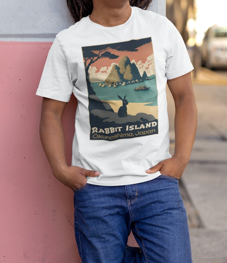 May include: White t-shirt with a vintage-style graphic of a rabbit island in Japan. The graphic features a small rabbit sitting on a rock, a boat in the water, and a mountain range in the background. The text "Rabbit Island" and "Okunoshima, Japan" is printed below the graphic.