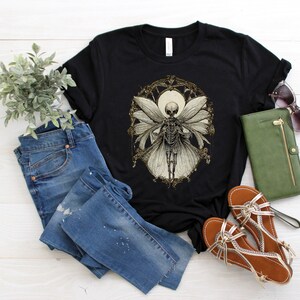 May include: Black t-shirt with a graphic of a skeleton fairy with wings, surrounded by a gold floral design. The shirt is laid out on a white wooden surface with blue jeans, a green wallet, and brown sandals.
