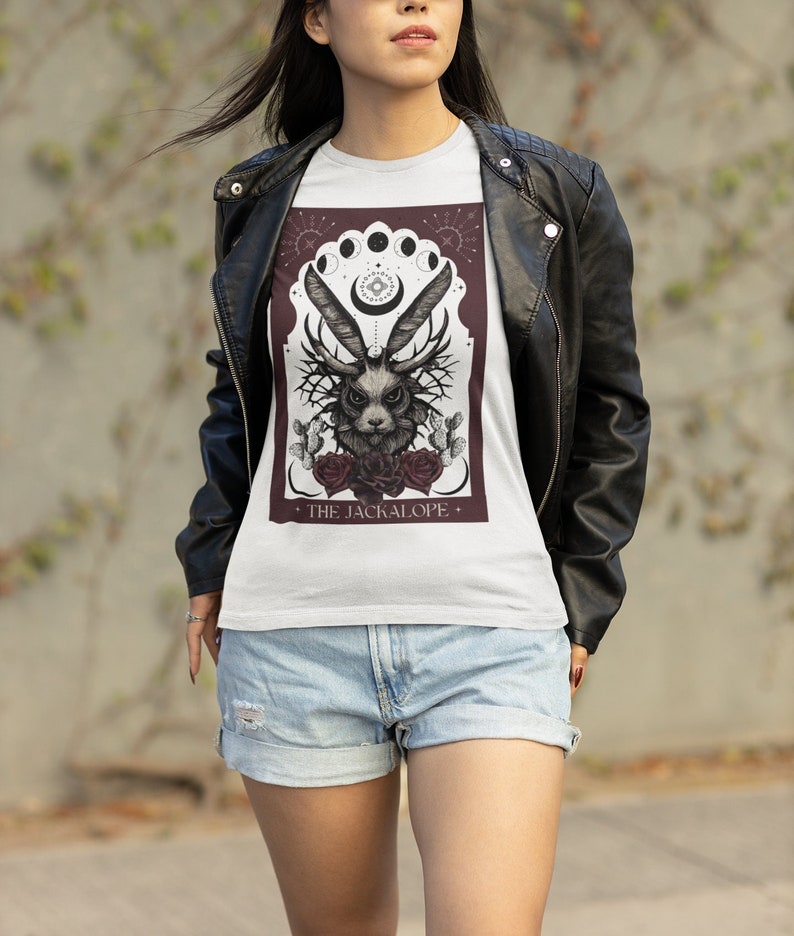 K&ouml;nnte beinhalten: Wei&szlig;es T-Shirt mit einer detaillierten Illustration eines Jackalopen mit Geweih, Rosen und Kakteen, umrahmt von einem burgunderfarbenen Bogen und Himmelselementen. Der Text "THE JACKALOPE" steht unten. Das Shirt wird mit Jeansshorts und einer schwarzen Lederjacke getragen.