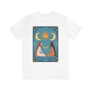 May include: White t-shirt with a graphic of the Lovers tarot card. The card features two women facing each other, a sun and moon, and the text "THE LOVERS".