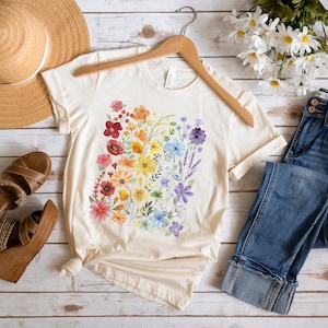 Puede incluir: Una camiseta de color crema con un dise&ntilde;o floral arco&iacute;ris. La camiseta est&aacute; colocada sobre una superficie de madera blanca con un sombrero de paja, un par de sandalias marrones, unos vaqueros azules y un ramo de flores blancas.