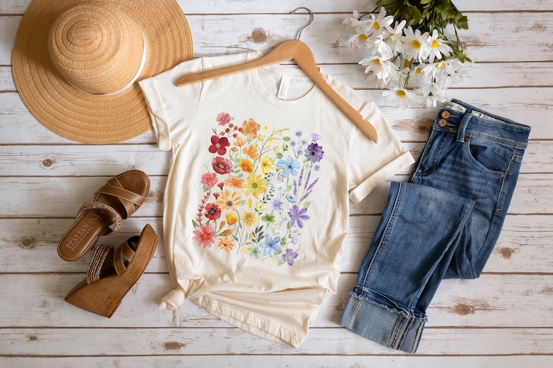 Puede incluir: Una camiseta de color crema con un dise&ntilde;o floral arco&iacute;ris. La camiseta est&aacute; colocada sobre una superficie de madera blanca con un sombrero de paja, un par de sandalias marrones, unos vaqueros azules y un ramo de flores blancas.