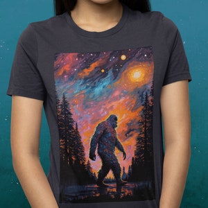May include: A dark grey t-shirt with a colorful graphic print of a Bigfoot walking through a forest under a starry sky.