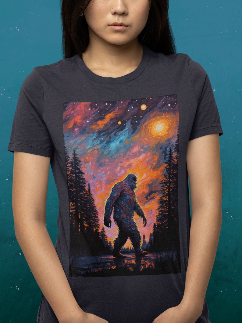 May include: A dark grey t-shirt with a colorful graphic print of a Bigfoot walking through a forest under a starry sky.