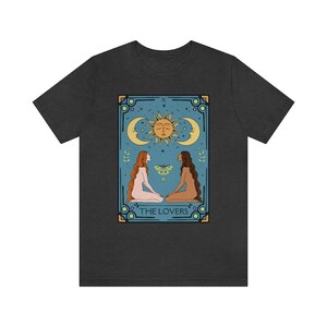 May include: A dark gray t-shirt with a graphic of the Lovers tarot card. The card features two figures, a sun, and a moon. The text "THE LOVERS" is printed below the figures.