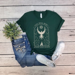 May include: A green t-shirt with a white graphic of a phoenix rising from flames. The graphic is surrounded by a floral design and the text "You will rise again and again".