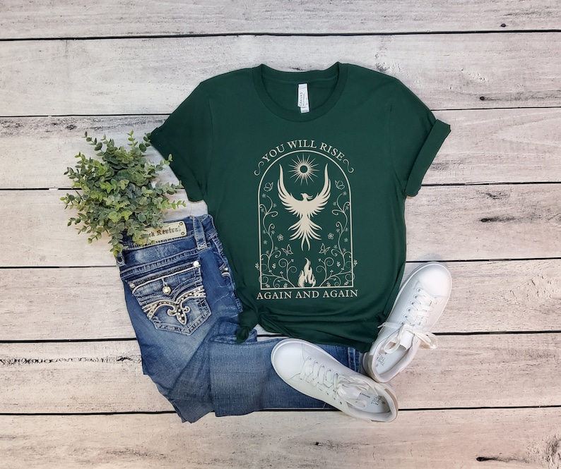 May include: A green t-shirt with a white graphic of a phoenix rising from flames. The graphic is surrounded by a floral design and the text "You will rise again and again".