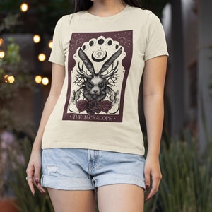 K&ouml;nnte beinhalten: Cremefarbenes T-Shirt mit detaillierter Grafik eines Jackalopen mit Geweih, Rosen und Kakteen, vor burgunderrotem Hintergrund mit Mondphasen. Der Text "THE JACKALOPE" befindet sich unten.