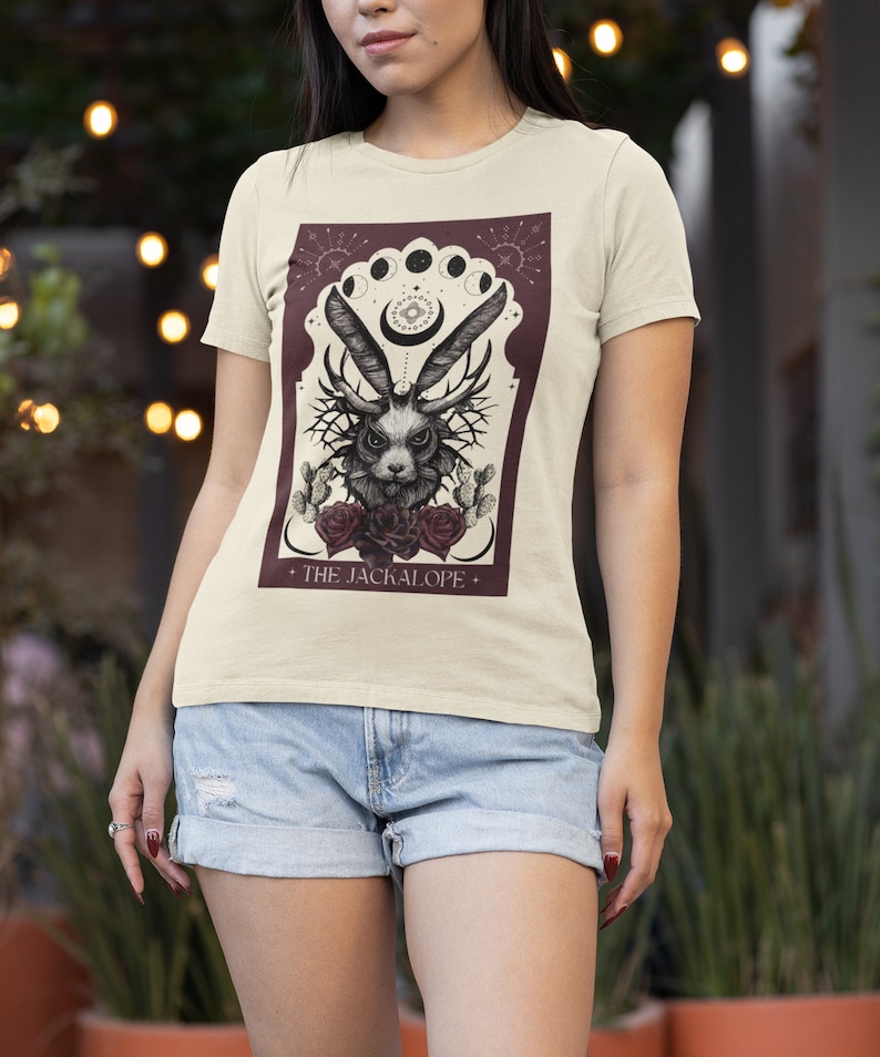 K&ouml;nnte beinhalten: Cremefarbenes T-Shirt mit detaillierter Grafik eines Jackalopen mit Geweih, Rosen und Kakteen, vor burgunderrotem Hintergrund mit Mondphasen. Der Text "THE JACKALOPE" befindet sich unten.