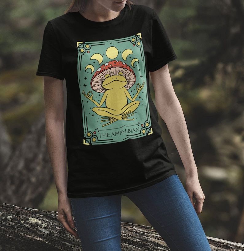 May include: Black t-shirt with a graphic of a green frog wearing a red and white mushroom hat. The graphic is surrounded by a gold frame and has the text "THE AMPHIBIAN" at the bottom.