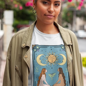 May include: A white t-shirt with a graphic of the Lovers tarot card. The card features two figures, a sun, a moon, and a butterfly. The text "THE LOVERS" is printed at the bottom of the card.