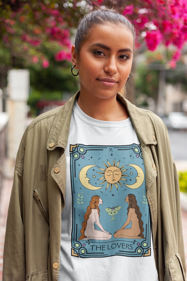 May include: A white t-shirt with a graphic of the Lovers tarot card. The card features two figures, a sun, a moon, and a butterfly. The text "THE LOVERS" is printed at the bottom of the card.