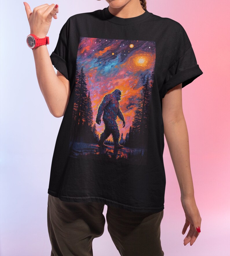 May include: Black t-shirt with a colorful graphic print of a Bigfoot walking through a forest under a starry sky.
