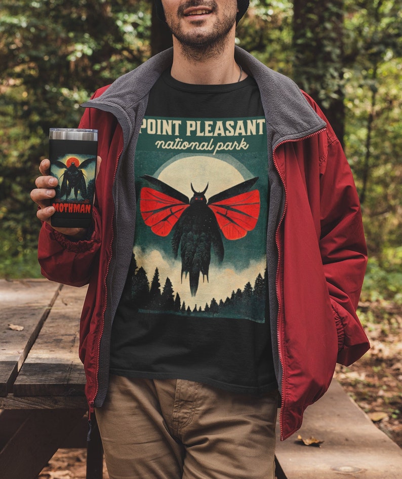 May include: A black t-shirt with a vintage-style graphic of a mothman with red wings. The graphic says "Point Pleasant National Park". A person is wearing the shirt and holding a black and silver travel mug with the words "Mothman" on it.