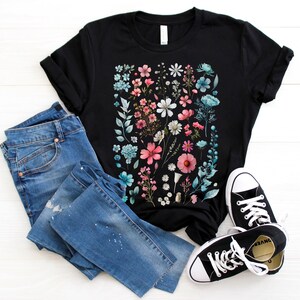 May include: Black t-shirt with a floral print featuring pink, blue, and white flowers on a black background. The shirt is paired with blue jeans and black sneakers.