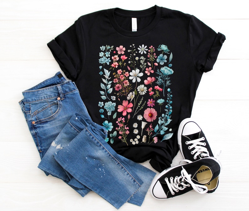 May include: Black t-shirt with a floral print featuring pink, blue, and white flowers on a black background. The shirt is paired with blue jeans and black sneakers.