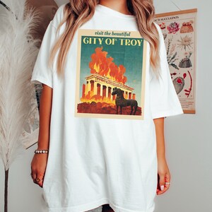 May include: White t-shirt with a vintage-style graphic of a burning building and a horse. The graphic says "visit the beautiful CITY OF TROY".