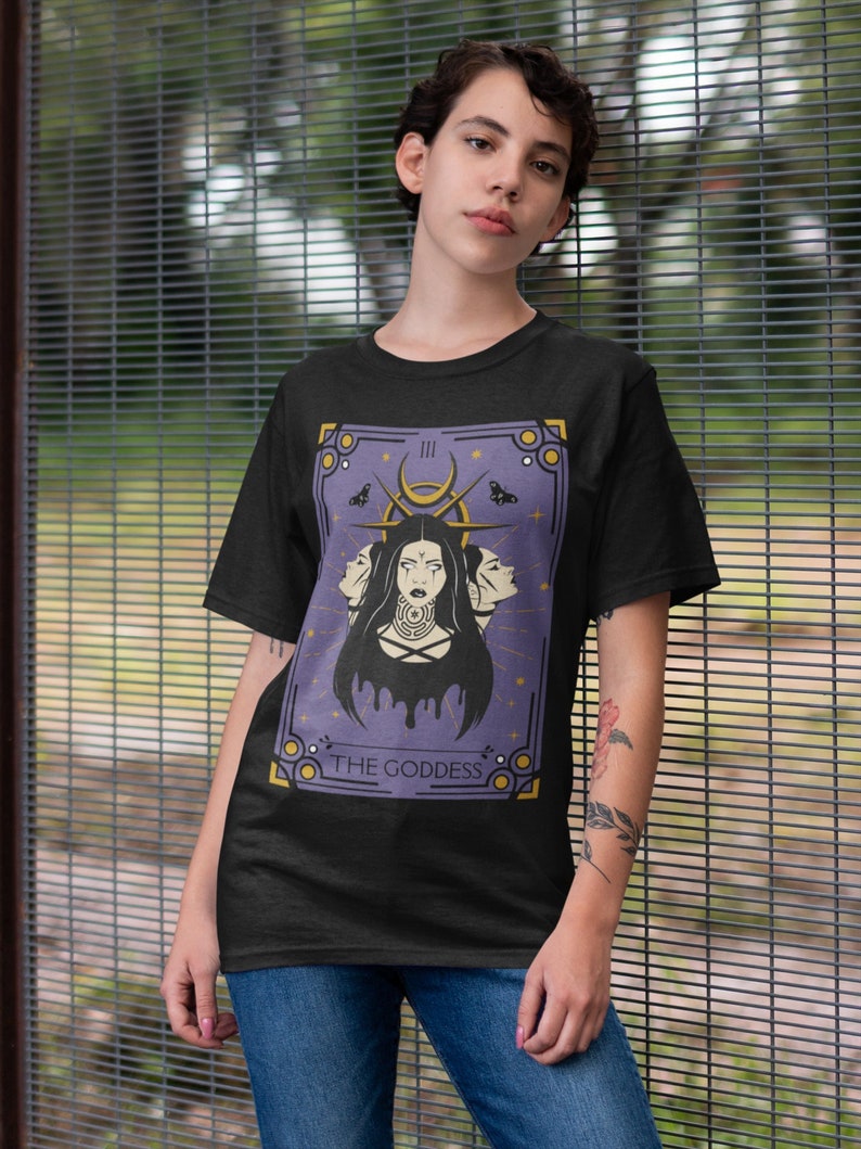 May include: A black t-shirt with a purple and gold illustration of a tarot card. The card is labeled "The Goddess" and features a woman with three faces.