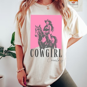 May include: A white t-shirt with a pink square featuring a silhouette of a cowgirl riding a horse. The text "COWGIRL Country" is printed below the image.