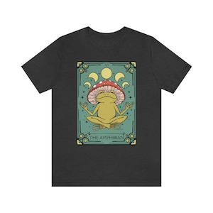 May include: Dark grey t-shirt with a green, yellow, and red illustration of a frog wearing a mushroom on its head. The frog is sitting in a meditative pose with its hands in a mudra. The illustration is surrounded by a decorative frame with the text "VI THE AMPHIBIAN".