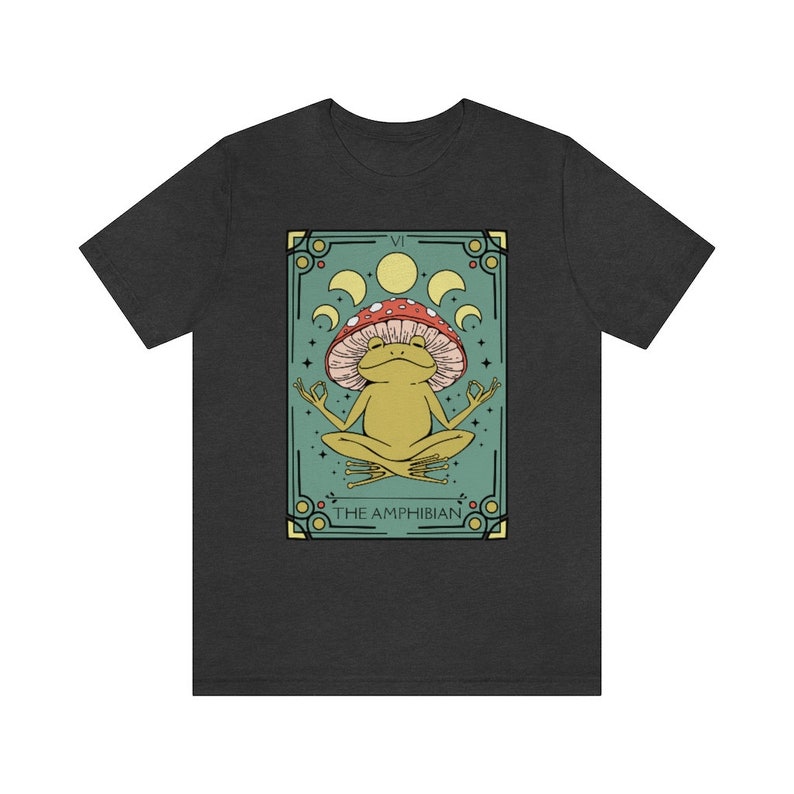 May include: Dark grey t-shirt with a green, yellow, and red illustration of a frog wearing a mushroom on its head. The frog is sitting in a meditative pose with its hands in a mudra. The illustration is surrounded by a decorative frame with the text "VI THE AMPHIBIAN".