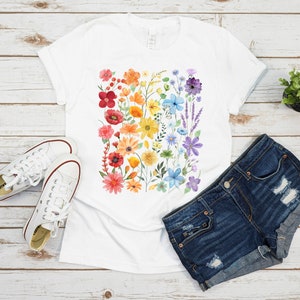 Puede incluir: Una camiseta blanca con un dise&ntilde;o floral arco&iacute;ris. Las flores est&aacute;n pintadas en acuarela y dispuestas en una franja vertical. La camiseta se combina con pantalones cortos de mezclilla y zapatillas blancas.
